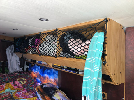 Inside an RV rental: view of a clever overhead storage net filled with essentials above a cozy bed and books.