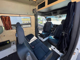 Interior view of an rv rental van, showing front and rear black cloth seating, light wood paneling, and a mounted fan.