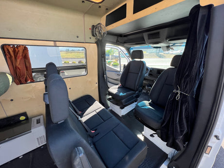 Interior view of an rv rental van, showing front and rear black cloth seating, light wood paneling, and a mounted fan.
