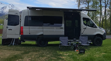 An RV rental camper van with open doors, awning, chairs, and table on grass against snowy mountains.