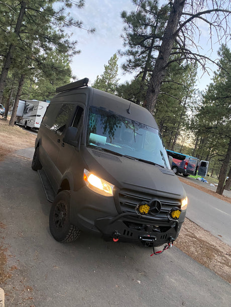 Dark gray Sprinter RV rental camper van with lights on, parked at a wooded campsite.