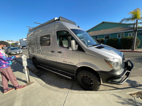 Shiny silver Winnebago Revel RV rental parked curbside on a sunny day, as a person operates a parking meter.