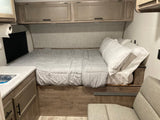 Cozy RV rental interior featuring a bed with light gray bedding, overhead cabinets, a small sink, and a cushioned seat.