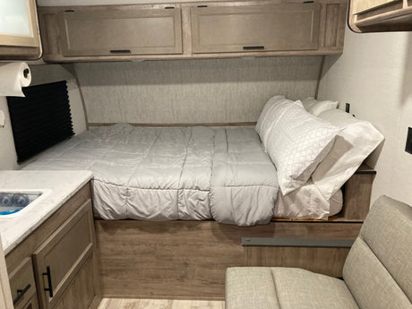 Cozy RV rental interior featuring a bed with light gray bedding, overhead cabinets, a small sink, and a cushioned seat.