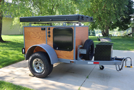  and front storage box.|Small custom camper trailer with a rooftop tent on a paved driveway. Ideal for RV rental guests seeking unique outdoor adventures.|Custom wood-paneled camping trailer with a rooftop tent and spare tire