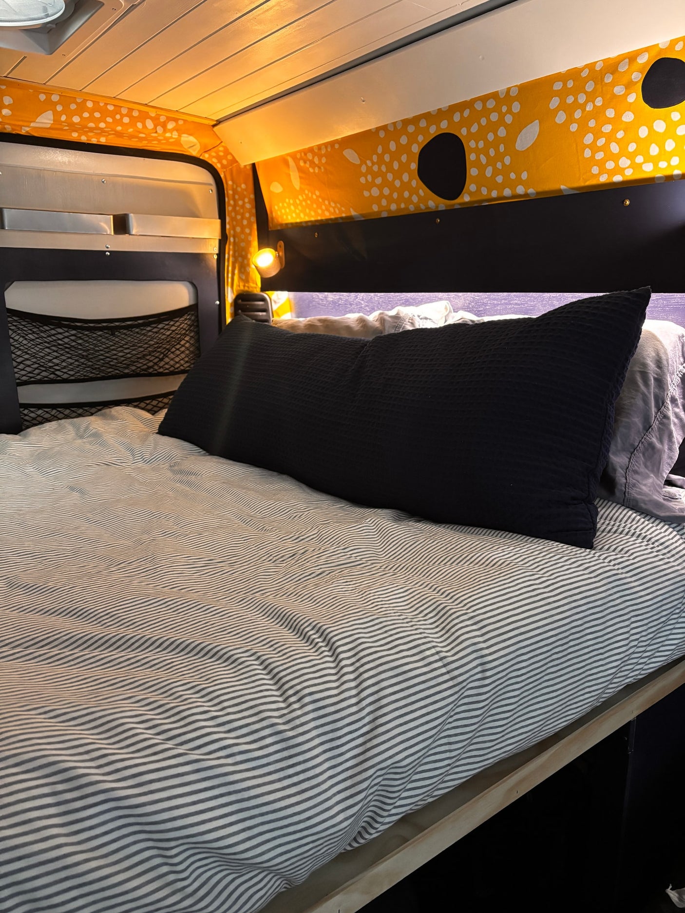 Cozy camper van RV rental bed with striped bedding, a black pillow, and bright yellow patterned upper wall trim.