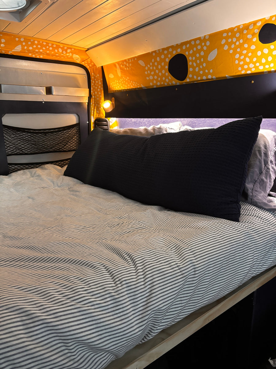 Cozy camper van RV rental bed with striped bedding, a black pillow, and bright yellow patterned upper wall trim.