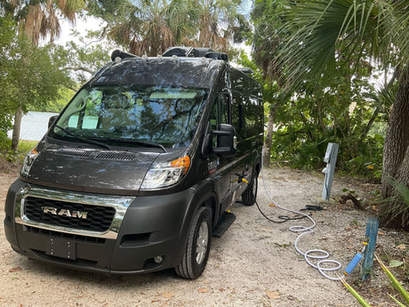 This dark gray camper van, an RV rental, is set up at a scenic campsite with power and water hookups by the water.