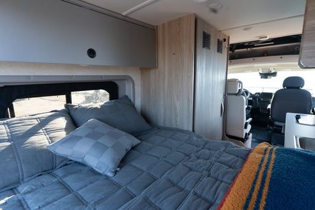 Cozy RV rental interior showcasing a comfortable bed with grey bedding, overhead storage, and the front cabin view.