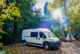 White camper van (RV rental) at a sunlit autumn forest campsite with other tents visible.