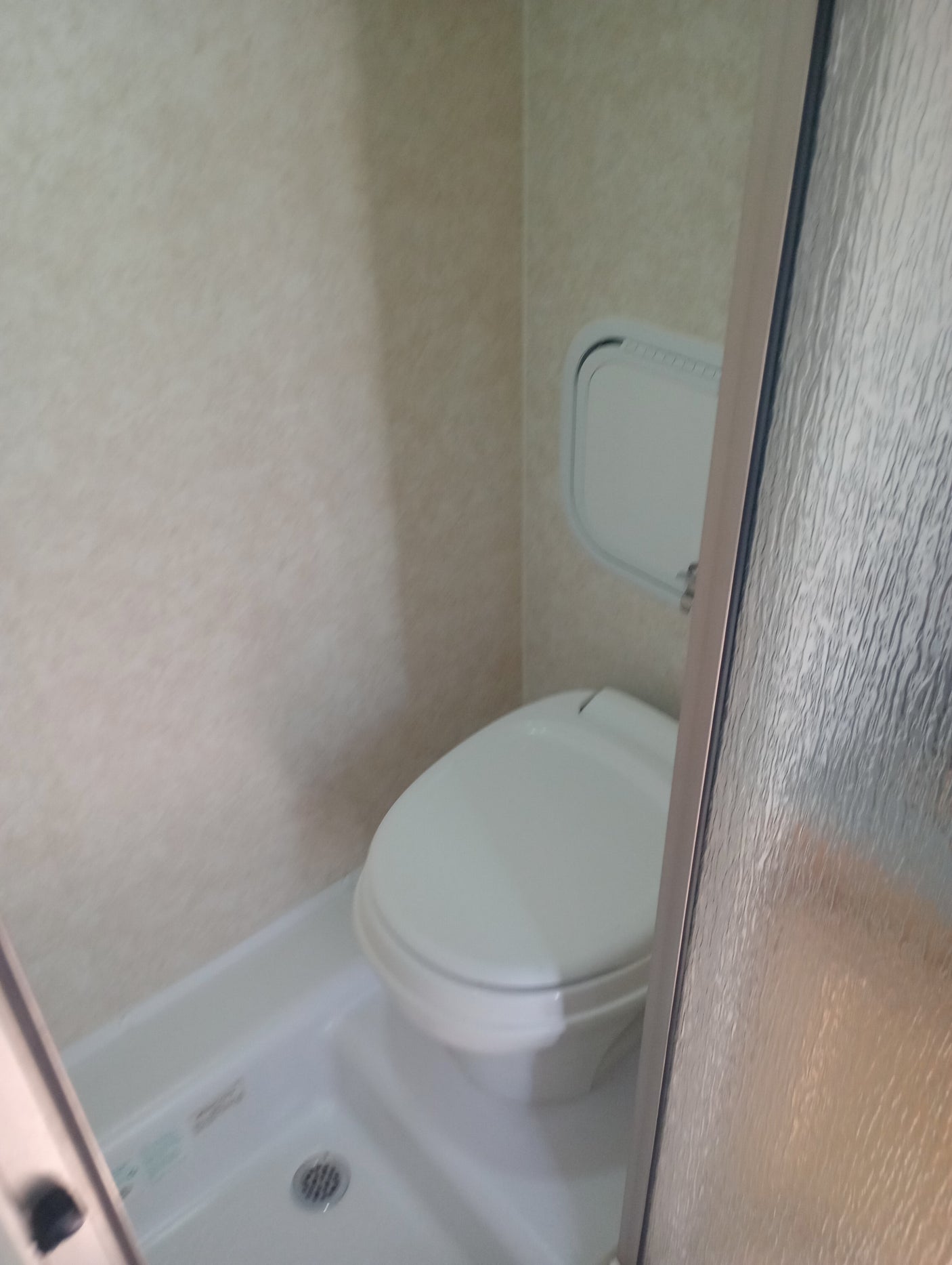 Compact RV rental bathroom featuring a toilet, shower, small wall panel, and textured glass door.