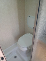 Compact RV rental bathroom featuring a toilet, shower, small wall panel, and textured glass door.