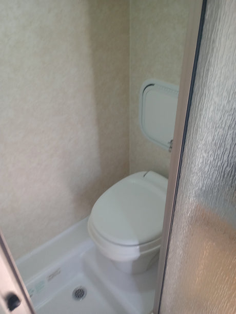 Compact RV rental bathroom featuring a toilet, shower, small wall panel, and textured glass door.