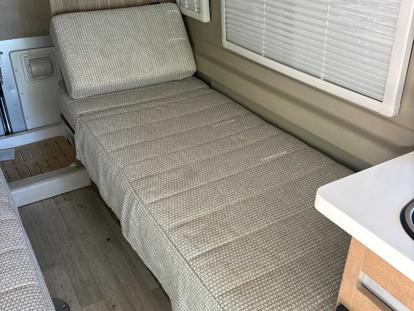 Inside an RV rental: a compact sleeping area with textured cushions, window blinds, and light wood flooring.