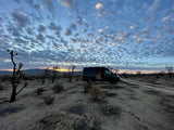 An RV rental parked in the desert at sunset, surrounded by Joshua trees under patterned clouds.