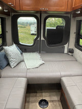 Cozy RV rental interior: rear seating/bed with pillows & blanket, wooden cabinets, and a field view from the back.