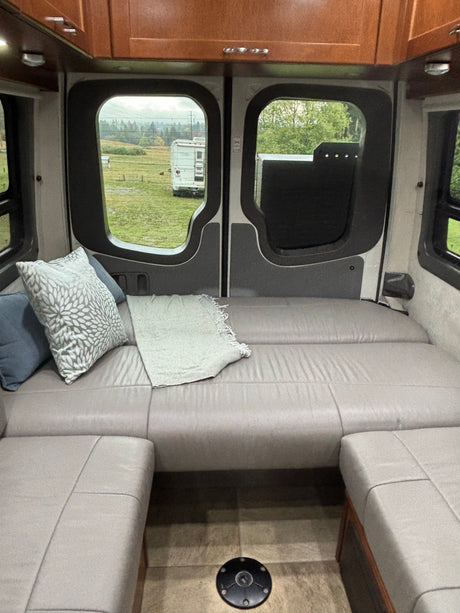 Cozy RV rental interior: rear seating/bed with pillows & blanket, wooden cabinets, and a field view from the back.