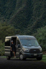 Man smiles from his grey RV rental van, parked amidst lush green mountains. Perfect for nature travel.