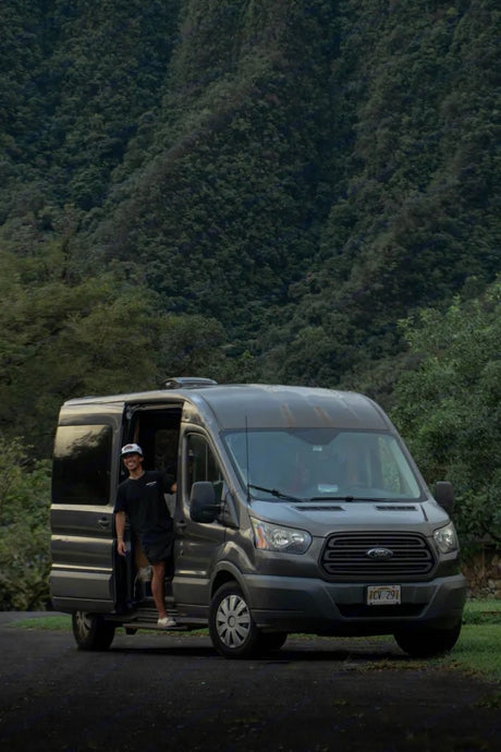 Man smiles from his grey RV rental van, parked amidst lush green mountains. Perfect for nature travel.