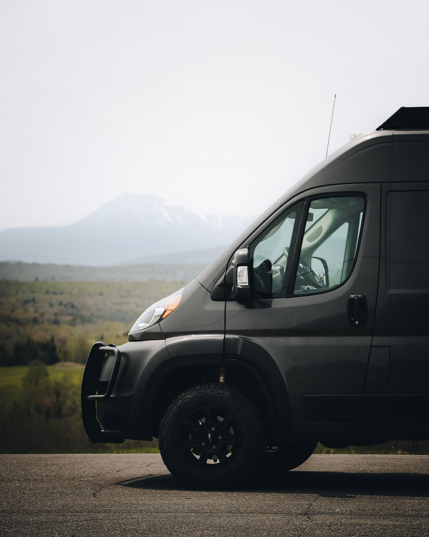 Adventure-ready dark grey RV rental van, with off-road tires and bull bar, parked on a road overlooking mountains.