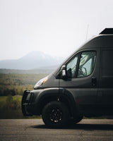 Adventure-ready dark grey RV rental van, with off-road tires and bull bar, parked on a road overlooking mountains.