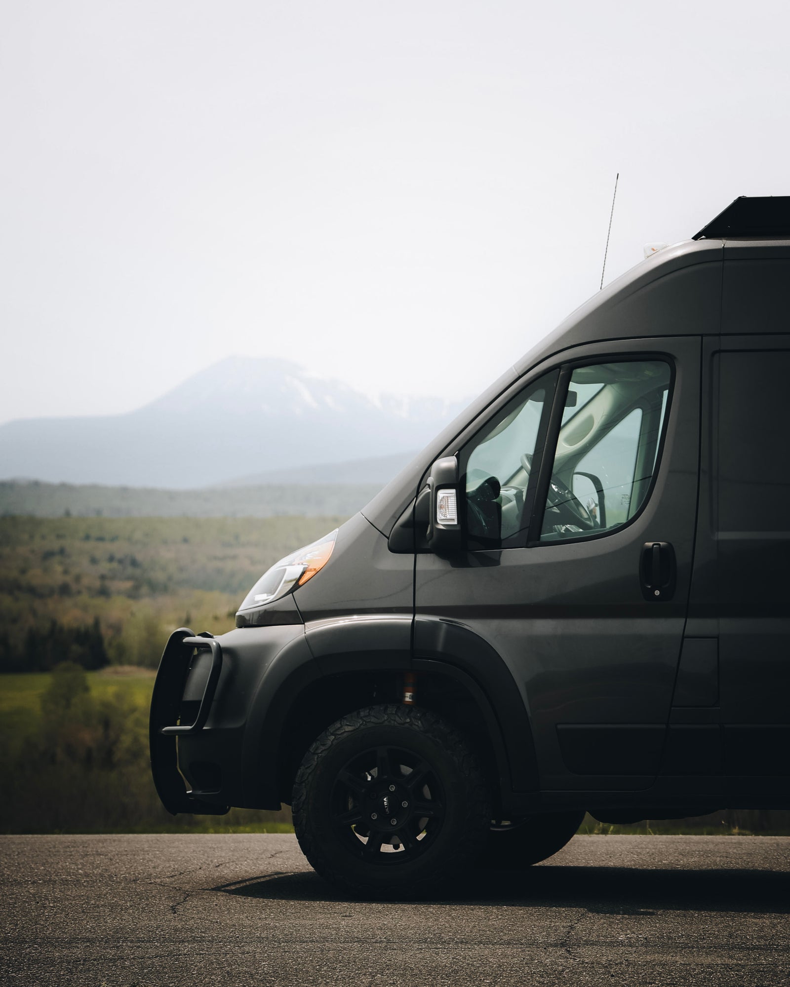 Adventure-ready dark grey RV rental van, with off-road tires and bull bar, parked on a road overlooking mountains.