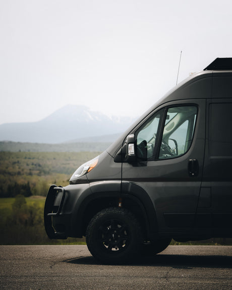 Adventure-ready dark grey RV rental van, with off-road tires and bull bar, parked on a road overlooking mountains.