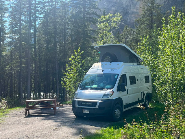 White RAM camper van (rv rental) with pop-top roof at a forest campsite with picnic table