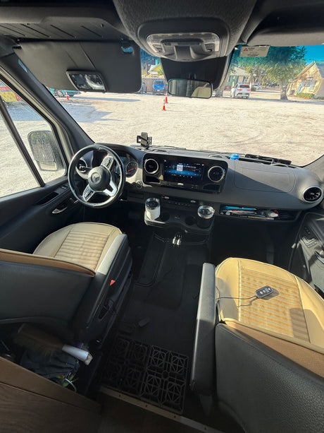 Spacious front cabin of an RV rental, featuring a sleek dashboard, steering wheel, and tan seats, parked on gravel.
