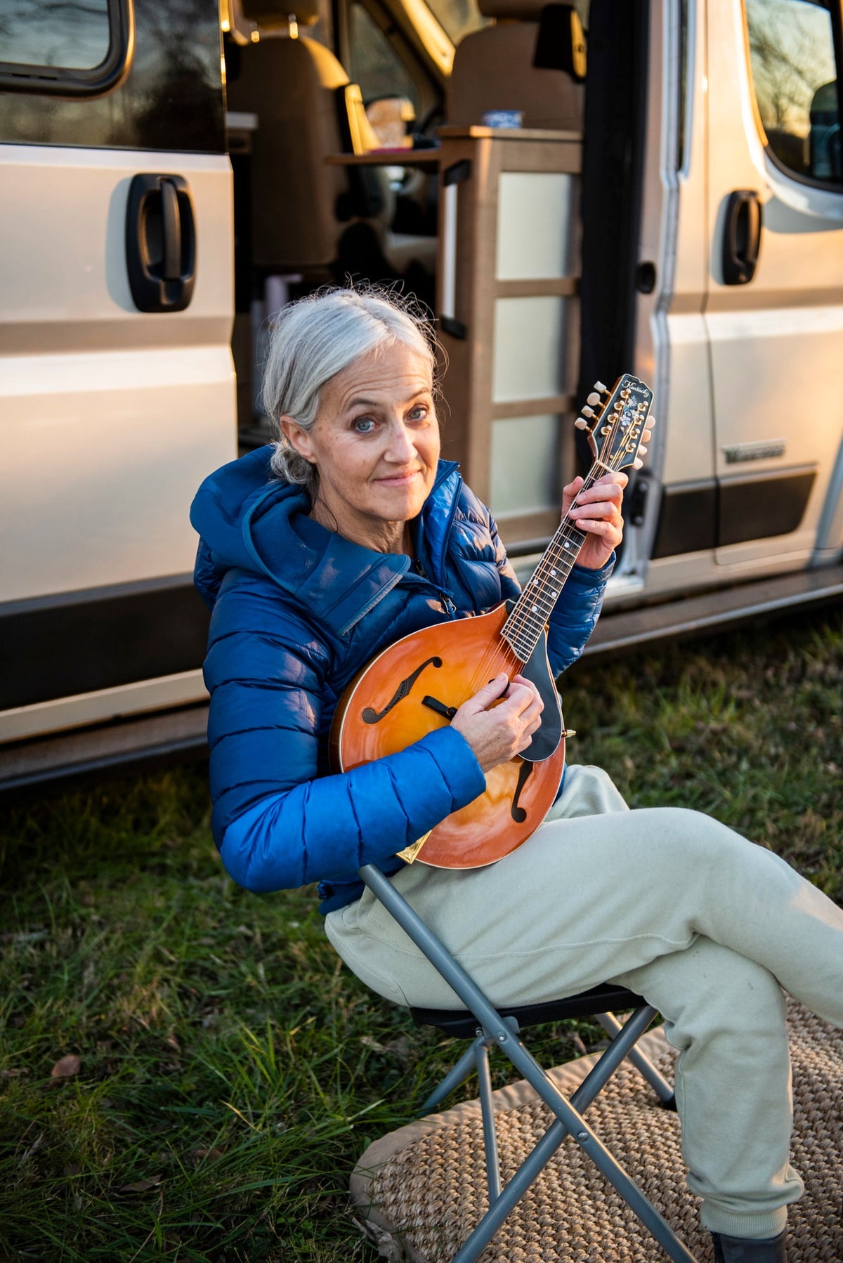 This smiling woman enjoys playing her mandolin outside her RV rental, sitting on a chair by the open camper van door. — photo 15