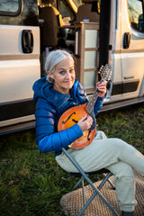 This smiling woman enjoys playing her mandolin outside her RV rental, sitting on a chair by the open camper van door.