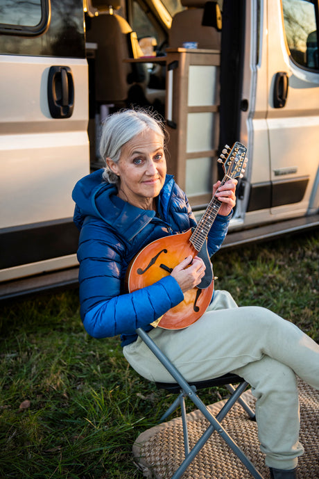 This smiling woman enjoys playing her mandolin outside her RV rental, sitting on a chair by the open camper van door.