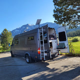 Scenic rv rental setup: gray camper van with open doors, ladder, Starlink, mountains, trees, and outdoor cooking.