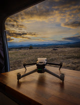 A drone on an RV's wooden table, framed by the window, overlooks a vibrant sunset landscape. Ideal for rv rental travel.