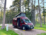This red **RV rental** is perfectly leveled at a tranquil pine forest campsite, pop-top extended for adventure.
