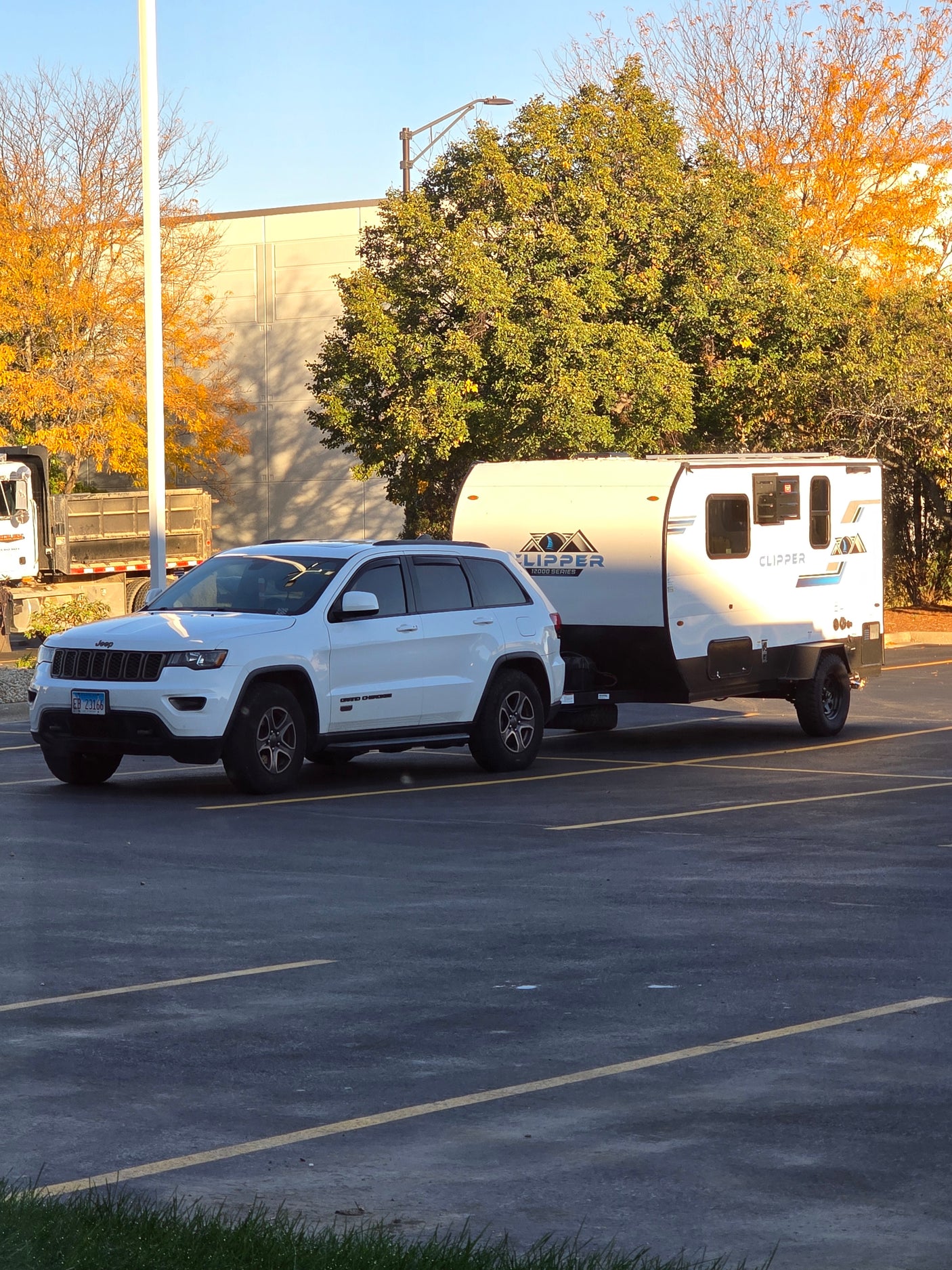 White Jeep Grand Cherokee towing a Clipper RV rental in a sunny parking lot, surrounded by autumn trees and a building.