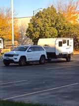 White Jeep Grand Cherokee towing a Clipper RV rental in a sunny parking lot, surrounded by autumn trees and a building.