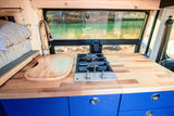 This RV rental features a compact kitchen with a wooden countertop, built-in stove, sink, and blue cabinets, offering a scenic water view.