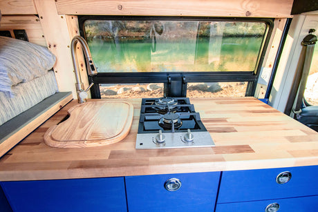This RV rental features a compact kitchen with a wooden countertop, built-in stove, sink, and blue cabinets, offering a scenic water view.