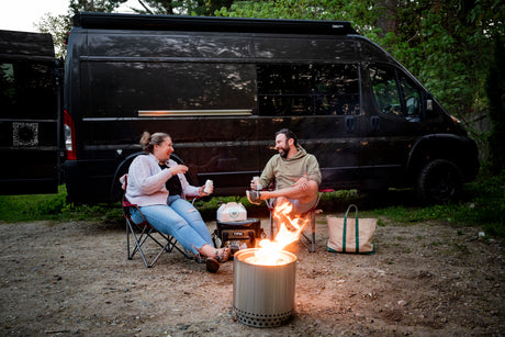 A happy couple in chairs by a Solo Stove fire pit and a black camper van, enjoying their RV rental experience.