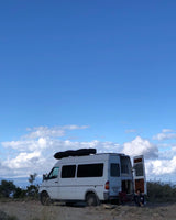 A white camper van, ideal for an RV rental, is parked on a scenic mountain road with open back doors and a person relaxing outside.