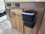 Functional RV rental interior with a hanging waste bin, power outlets, and light switches on wood grain cabinetry.