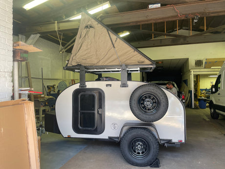 A light gray teardrop camper with a deployed rooftop tent, ideal for an RV rental. Parked in a workshop.