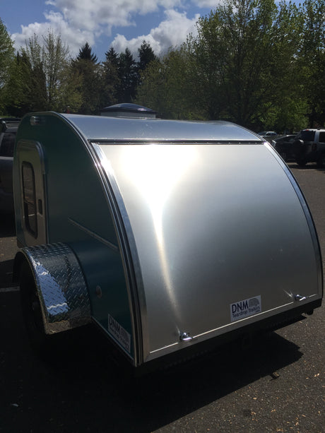 A stylish silver and teal teardrop trailer parked outdoors, reflecting sunlight. Consider this compact rv rental for your next trip.