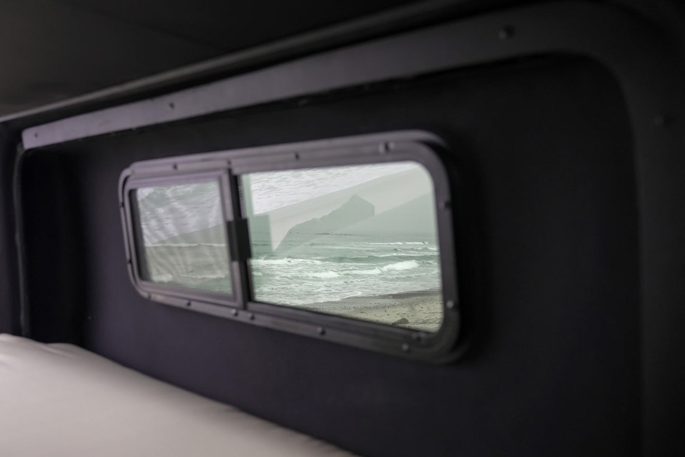 Cozy RV rental interior, window offers ocean beach view with waves & distant bluff. Ideal for travel.