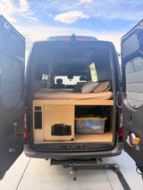 RV rental camper van interior. Features a comfortable bed, custom wood storage, and open rear doors, ideal for mobile living.