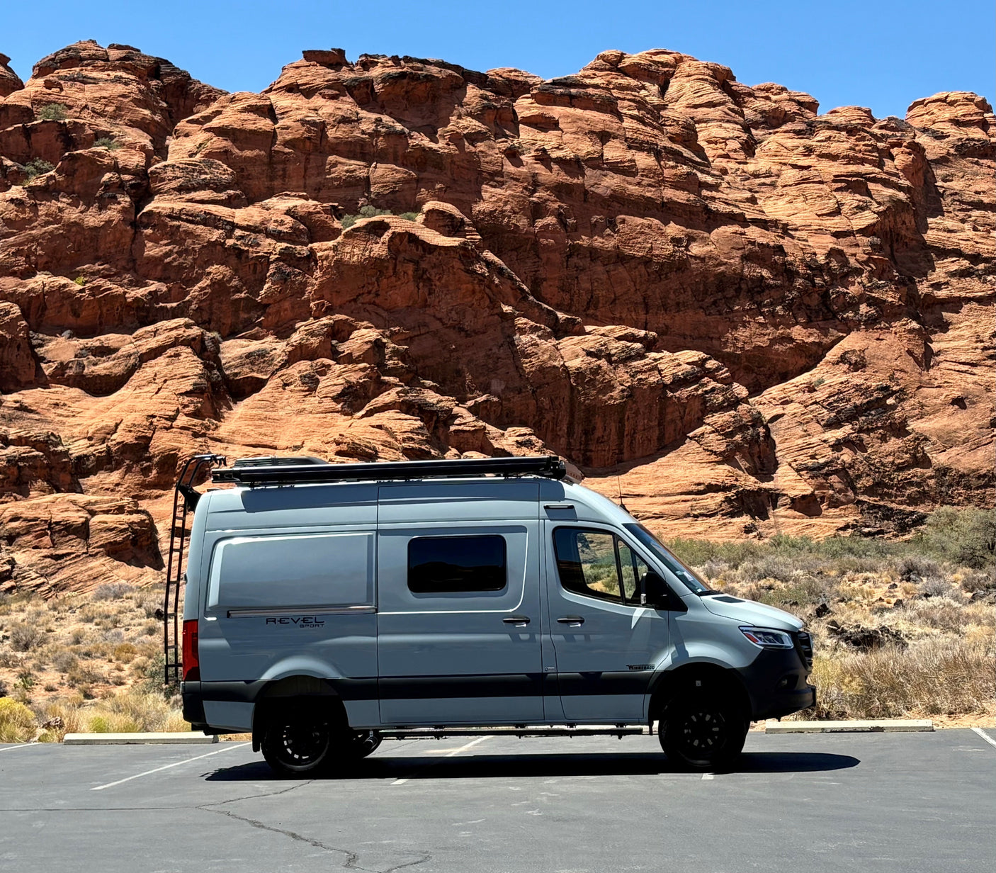 A light gray Winnebago Revel camper van, perfect for an RV rental, is parked in a desert landscape with dramatic red rock formations.