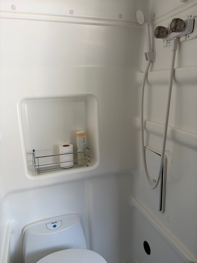 White RV rental bathroom featuring a compact toilet, handheld shower, and essentials on a shelf. — photo 3