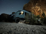 Cozy night camping! This RV rental, a Mercedes Sprinter van, glows under a starry sky next to a huge rock formation in the desert.