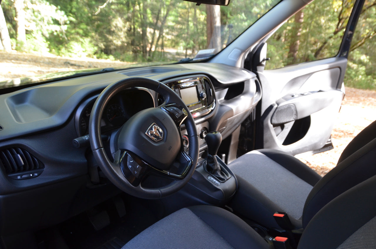 Inside an RV rental van: driver's view of dashboard, steering wheel (Ram logo), and manual transmission. Open door shows outdoor scenery.